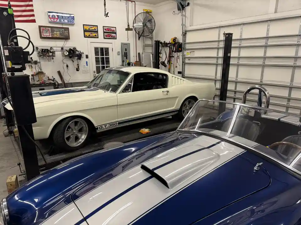 A white 1960s Ford Mustang Shelby GT350 is parked on a lift in a well-equipped garage, with a blue and white classic car in…