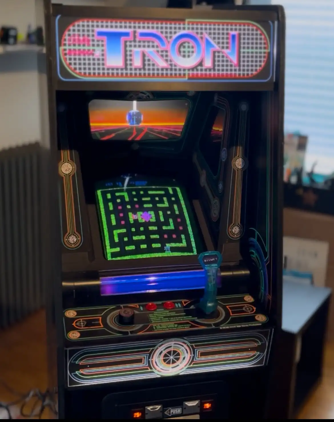 A vintage TRON arcade machine is shown with its screen displaying a glowing green maze and a blue light cycle, while the top…