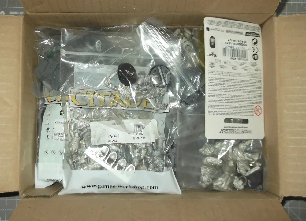 An open cardboard box contains several clear plastic bags filled with unpainted, silver-colored miniature figurines, likely…