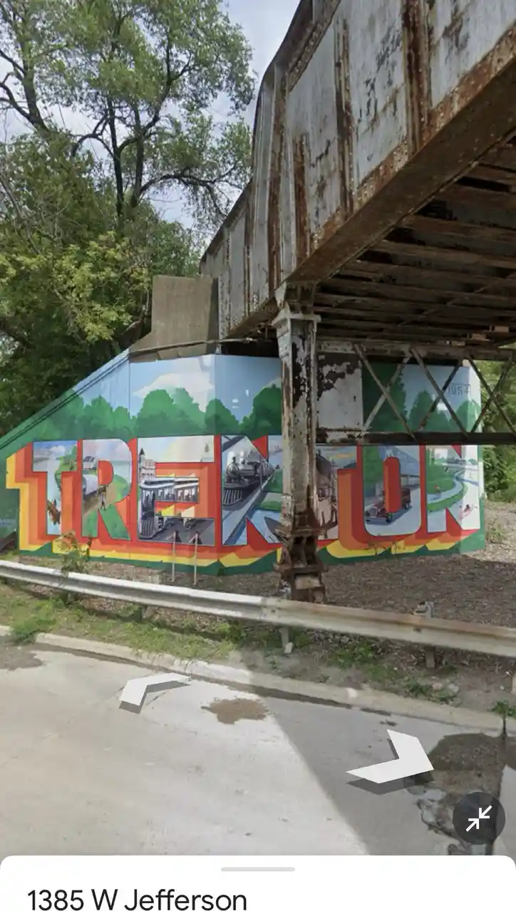 A Google Street View image shows a rusty metal railroad overpass crossing over a road with a large mural painted on the…