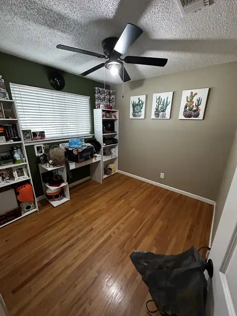 A tidy, well-lit room with hardwood floors, white shelves filled with books and decor, cactus wall art, and a ceiling fan…