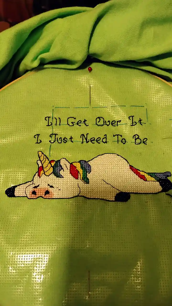 A close-up of a green fabric held in an embroidery hoop, showing a partially stitched cartoon unicorn lying down with a…