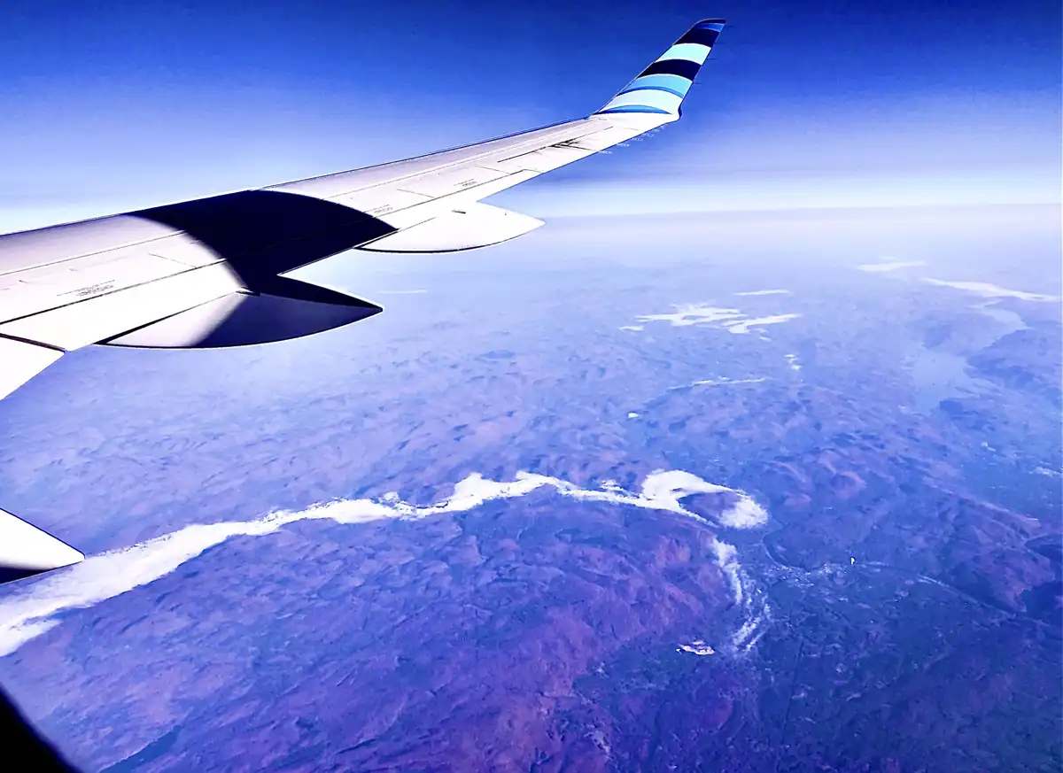 An aerial view from an airplane window shows the wing extending over a vast, hazy landscape with winding white rivers or…
