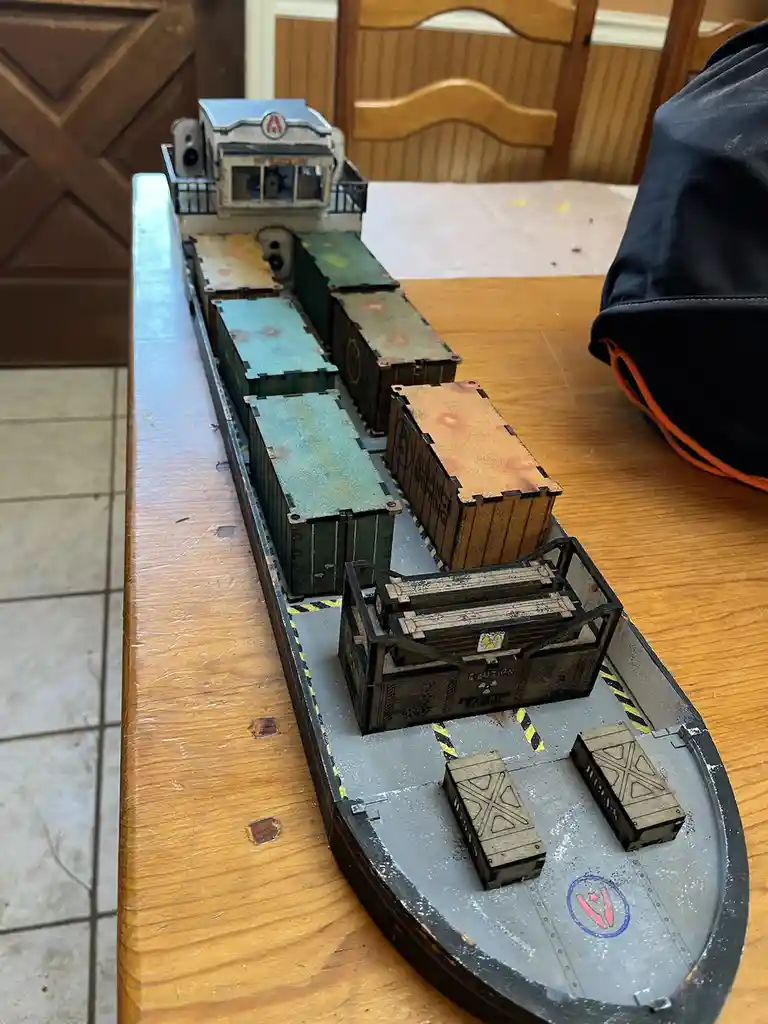 A detailed model ship, likely for a tabletop game, sits on a wooden table. The ship's deck is loaded with various painted…