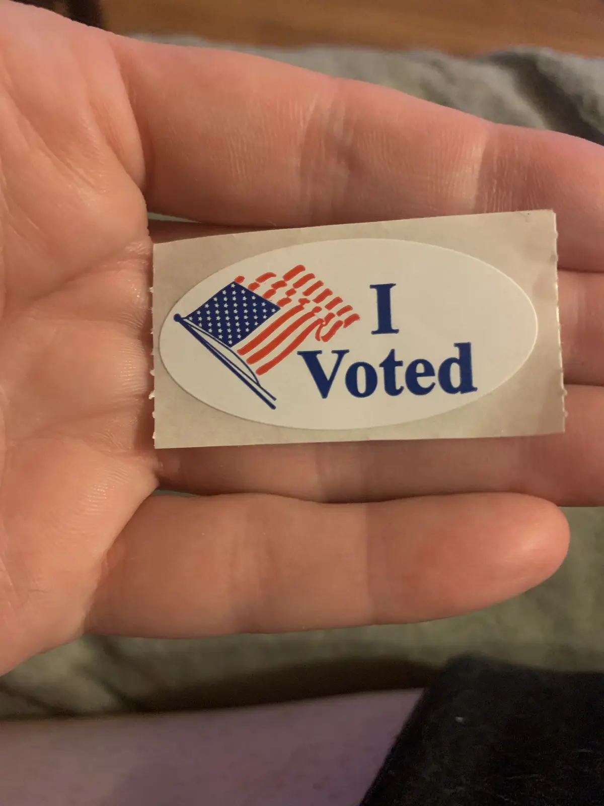 A hand holds a small, oval-shaped 'I Voted' sticker featuring a stylized American flag. The sticker is white with blue text…