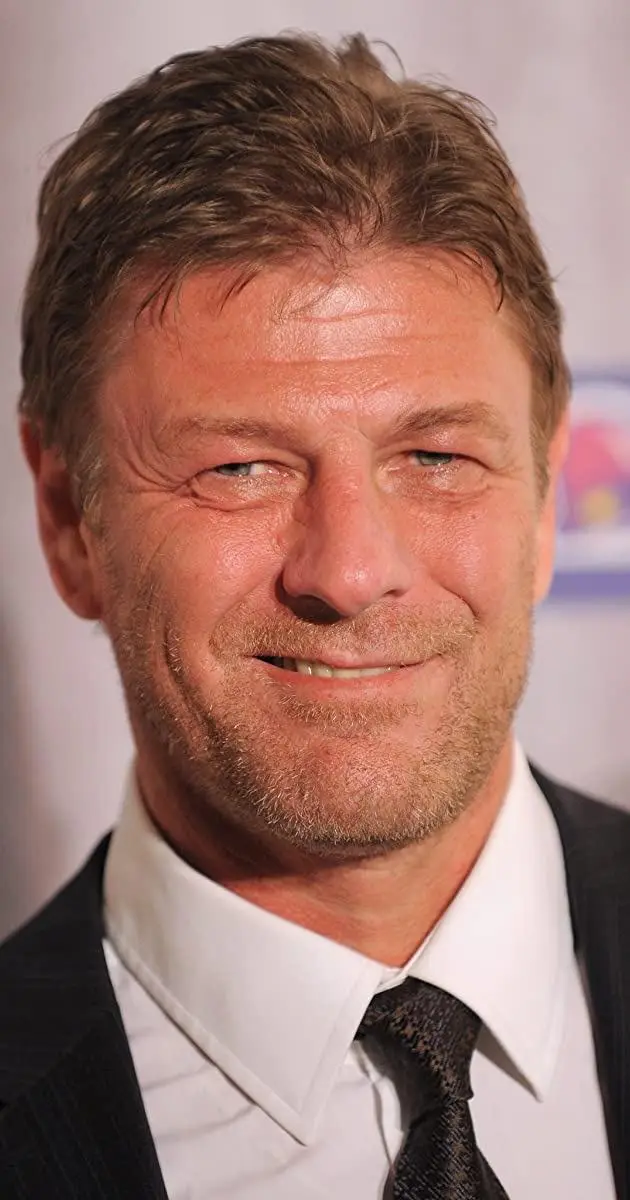 A close-up photo of a man with short brown hair, stubble, and a slight smile, wearing a dark suit, white shirt, and…