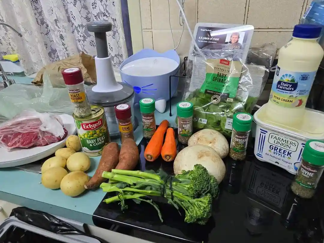 A kitchen counter is cluttered with groceries including raw meat, potatoes, sweet potatoes, carrots, broccoli, spinach…