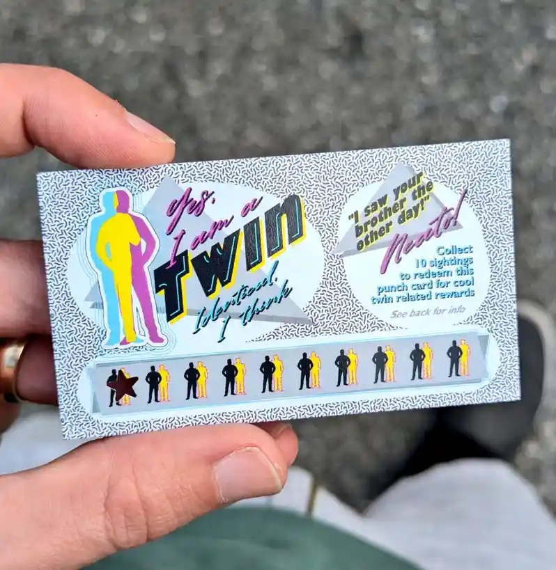 A hand holds a retro-style punch card with a 1980s aesthetic, featuring colorful twin silhouettes and text declaring 'Yes, I…