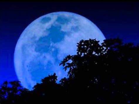 A large, vividly blue moon dominates a dark night sky, with the silhouettes of tree branches visible in the foreground. The…