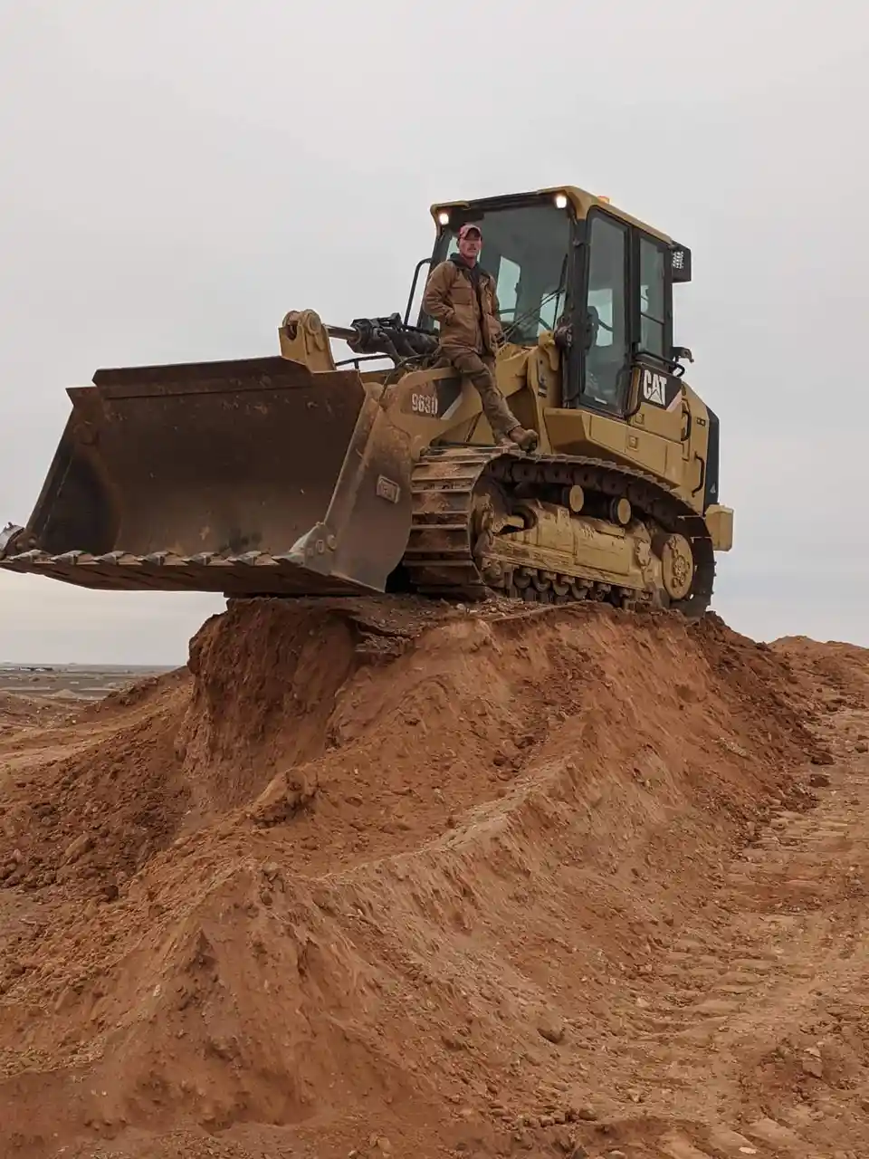 A man in work clothes and a cap is standing on the side of a yellow Caterpillar 963D bulldozer, which is perched…