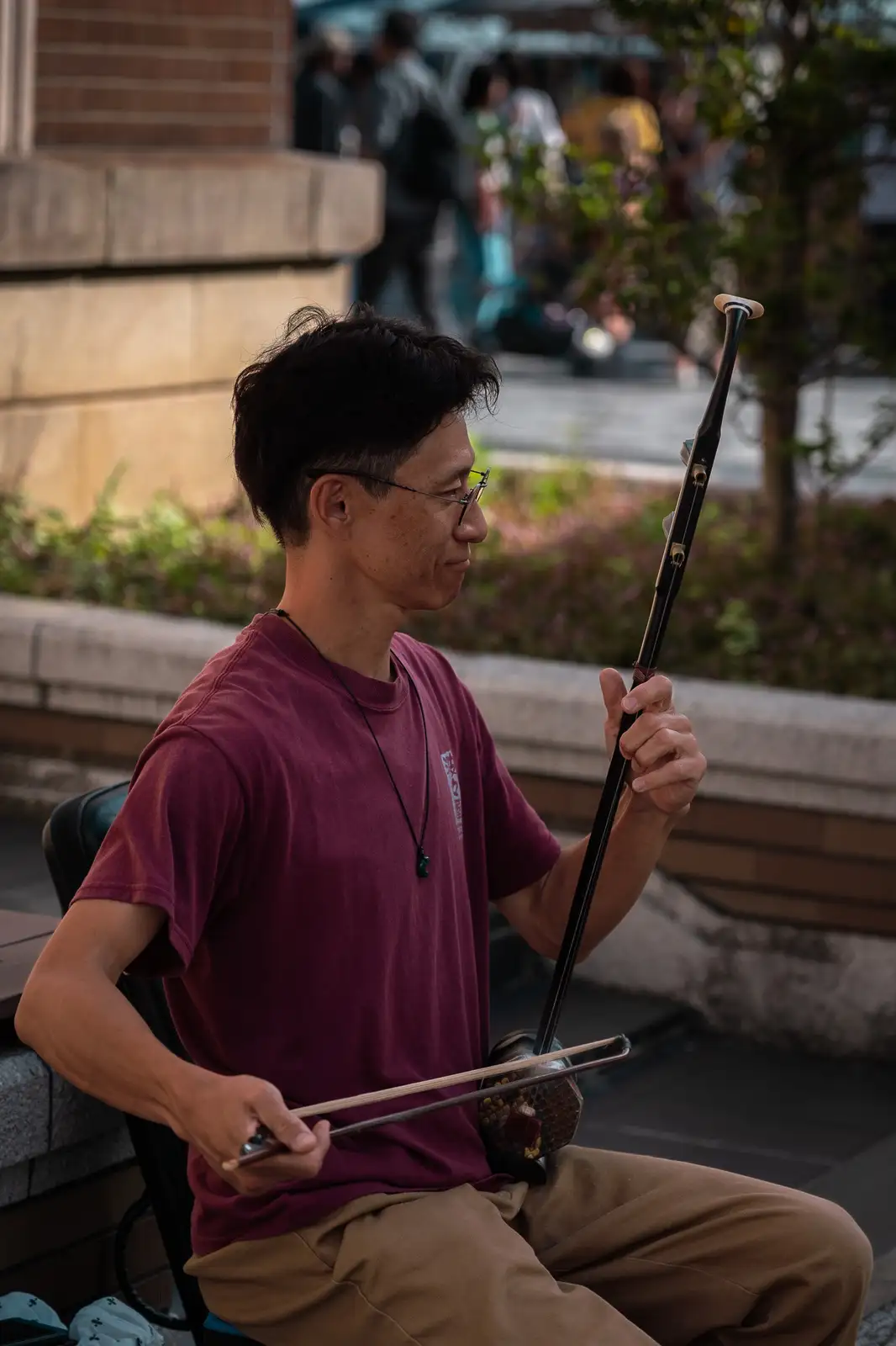 A man in a maroon t-shirt and glasses sits on a bench outdoors, holding a traditional Chinese two-stringed fiddle (erhu) and…