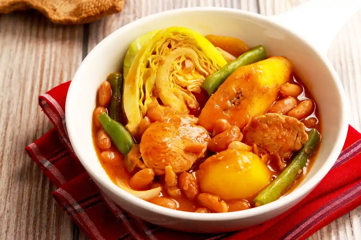 A white bowl filled with a hearty stew containing chicken, beans, green beans, cabbage, and potatoes, resting on a red plaid…