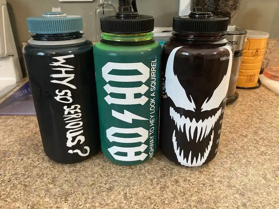 Three water bottles sit on a kitchen counter. The left bottle is black with white text reading 'WHY SO SERIOUS?'. The middle…