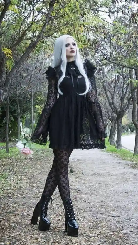 A person with long white hair and dark makeup stands on a leafy park path wearing a black lace dress, matching lace tights…