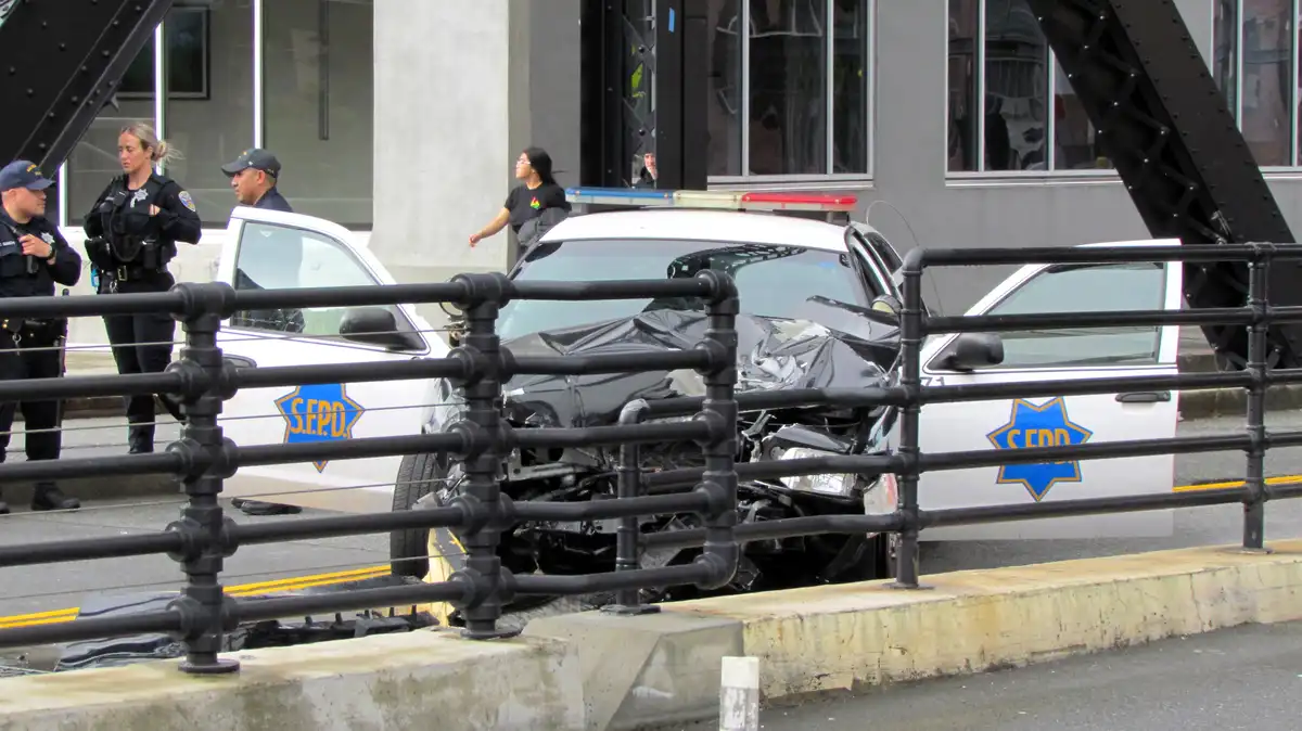 A severely damaged white S.F.P.D. police car is shown with its front end crushed and resting on a concrete barrier behind a…