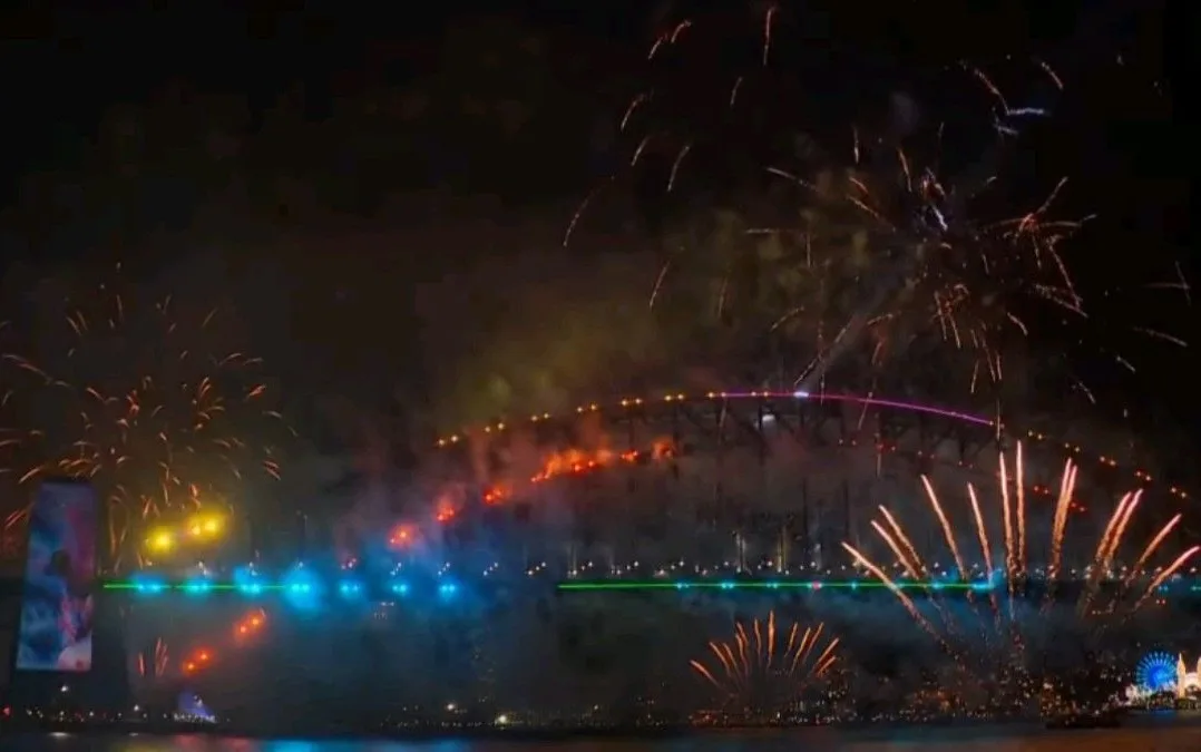 A nighttime view of the Sydney Harbour Bridge illuminated with colorful lights and surrounded by fireworks exploding in the…