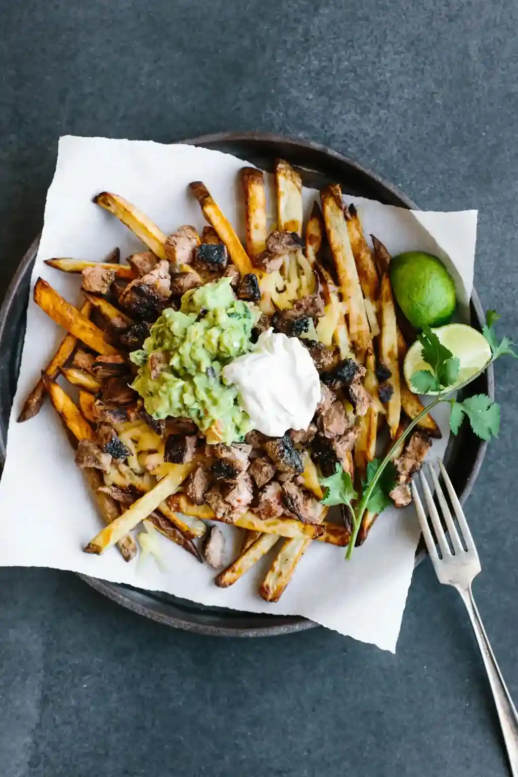An overhead shot of a plate of loaded fries topped with guacamole, sour cream, shredded cheese, and meat, garnished with…