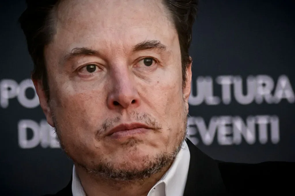 a close-up photo of elon musk with a serious, slightly annoyed expression, wearing a black suit and white shirt, against a…